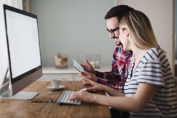 Eine Frau und ein Mann sitzen am Tisch vor einem Computer und gucken parallel auf ein Tablet. | © Adobe Stock