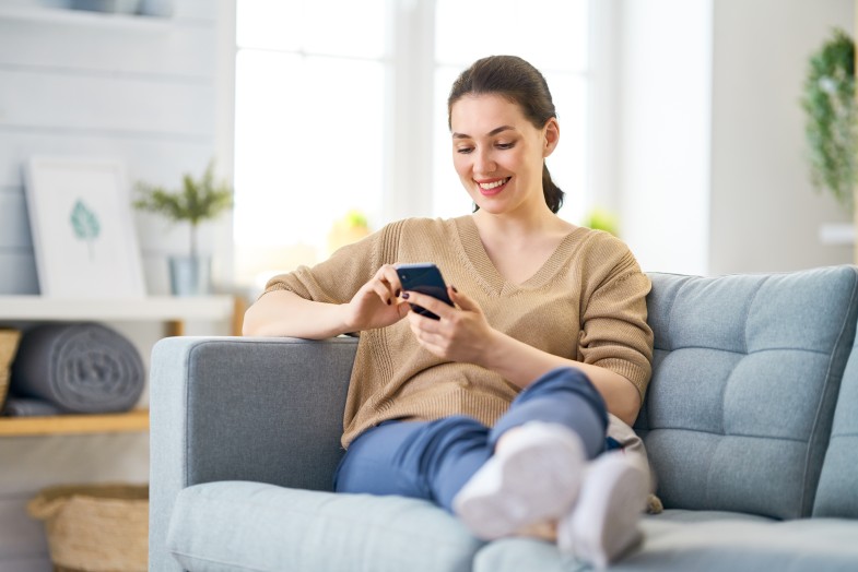Bild: Eine junge Frau sitzt auf dem Sofa. Sie lächelt. Sie hat ein Telefon in der Hand.