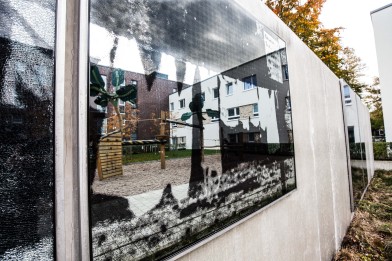 Sie sehen auf der großen Spiegelflächen einer Sichtschutzmauer das historische Foto einer Wäscheleine. Im Hintergrund sehen Sie durch die Spiegelwand ein Wohnquartier mit Spielplatz.